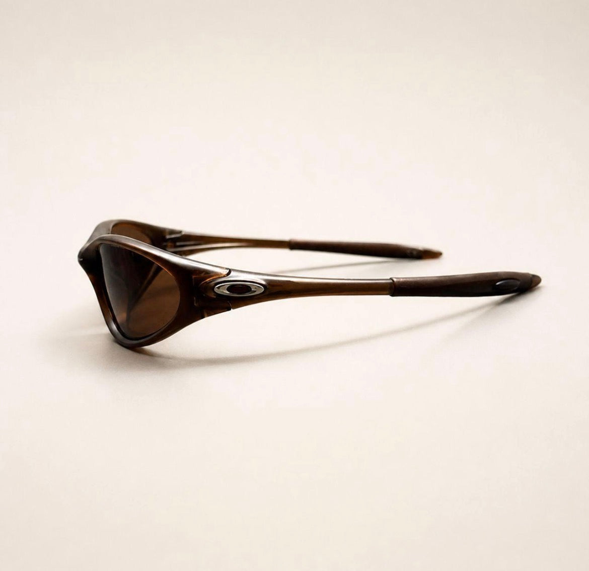 Side view of brown Oakley Minute sunglasses, showing the brown frame, lens and silver oval temple detail.
