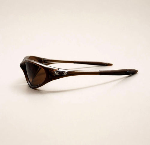 Side view of brown Oakley Minute sunglasses, showing the brown frame, lens and silver oval temple detail.